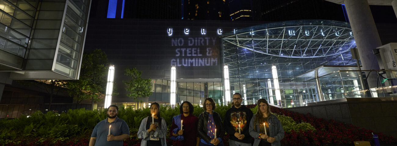 Candlelit vigil in front of GM's HQ with projections on dirty steel and aluminum behind.