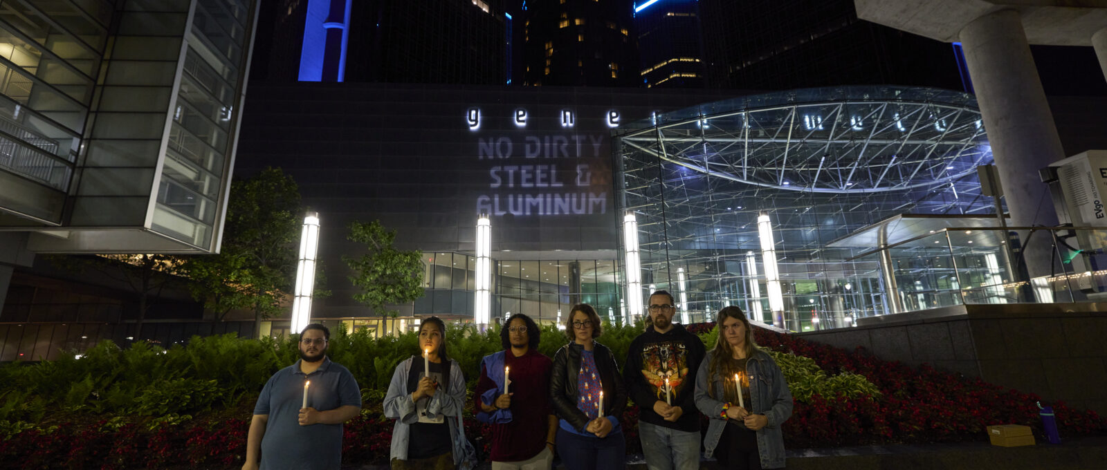 Candlelit vigil in front of GM's HQ with projections on dirty steel and aluminum behind.